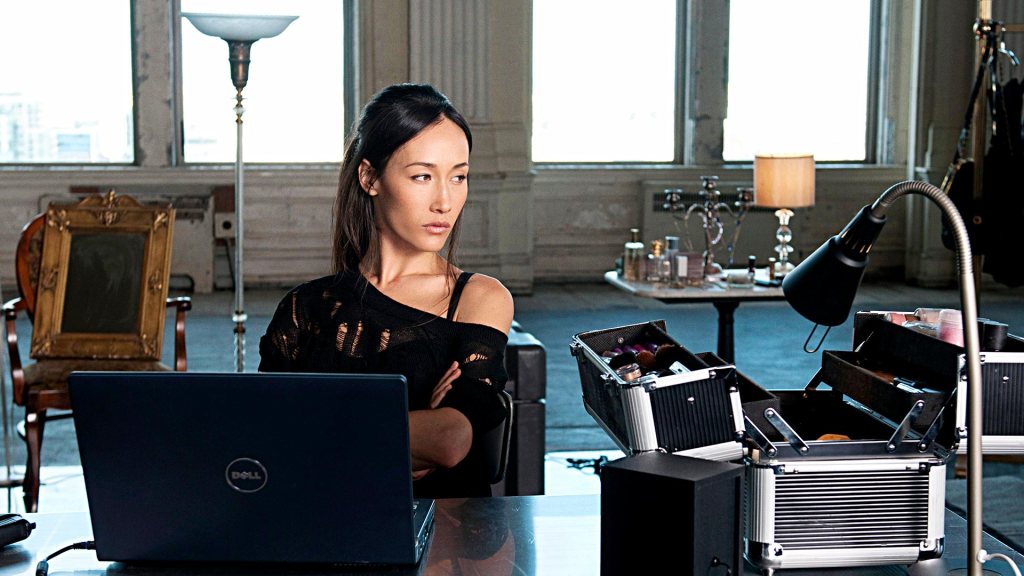 Nikita (played by Maggie Q) sitting in front of a desk with an open laptop , a lamp, and makeup kit bag on it