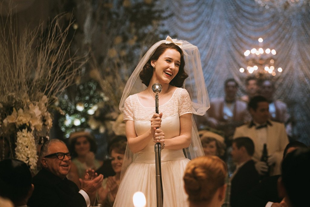 Midge (played by Rachel Brosnahan) wearing a wedding dress making a speech at her own wedding with her guests sitting at tables around her